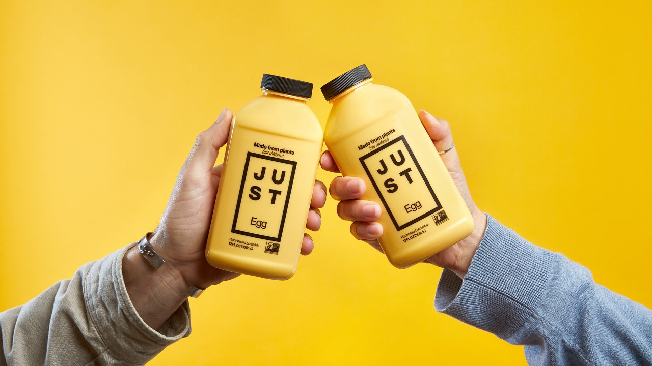 Produktfoto: Eat Just Två händer som håller upp en varsin plastflaska mot varandra mot en gul bakgrund.