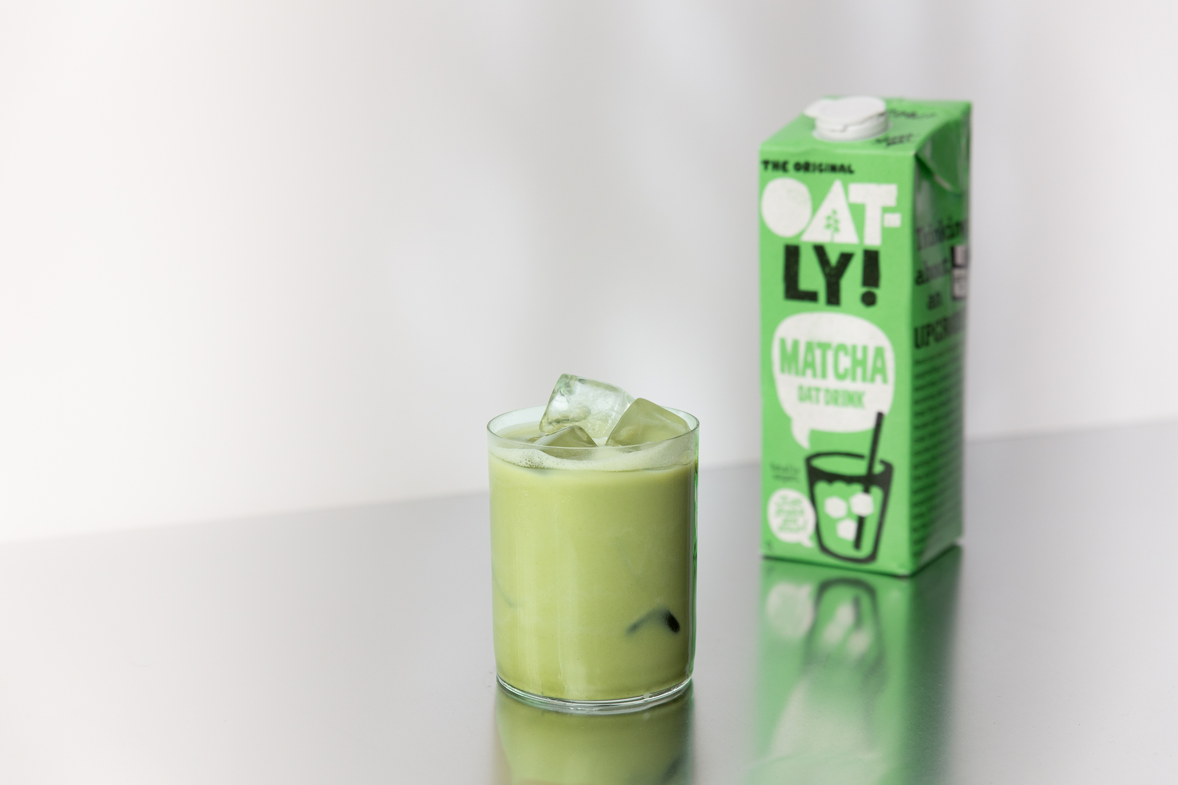 Produktfoto: Oatly Ett glas med matcha latte och en tetra med färdigblandad matcha latte bredvid på ett silverbord.