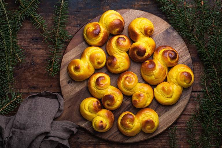 Freshly baked homemade Swedish traditional s-shaped saffron buns, also known as lussebullar or saffransbröd. Seen from above flat lay on a dark wooden table surrounded by spruce branches.