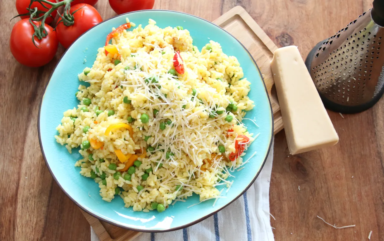 Risotto med gröna ärtor på en turkos tallrik, bredvid tallriken ligger en vegansk parmesanost och några tomater
