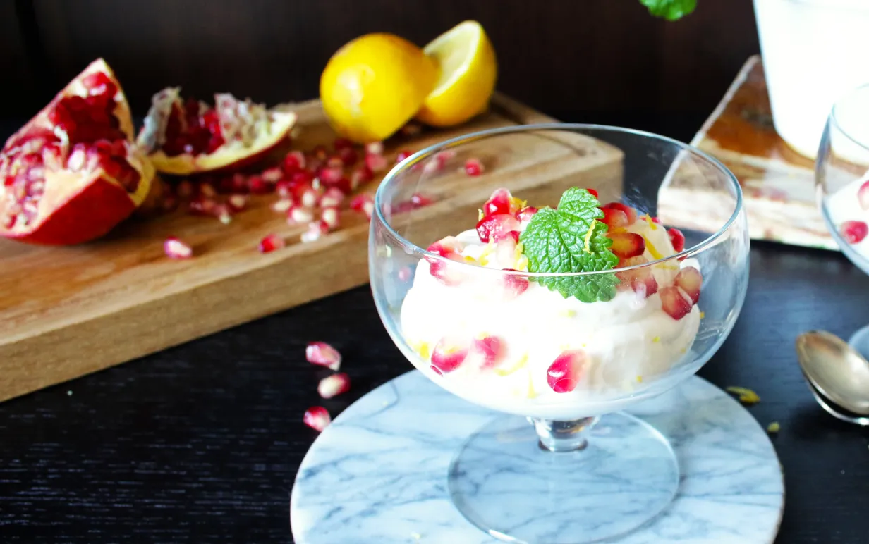 Glasskål med citronmousse och granatäppelkärnor framför en träskiva med granatäpplen och citroner.