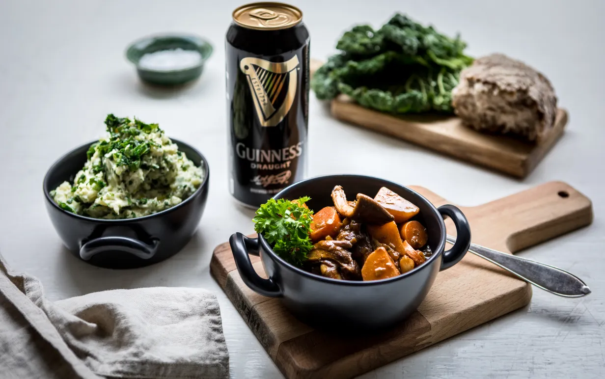 En skål med Irish stew står på en skärbräda bredvid en skål med irländskt potatismos och en öl