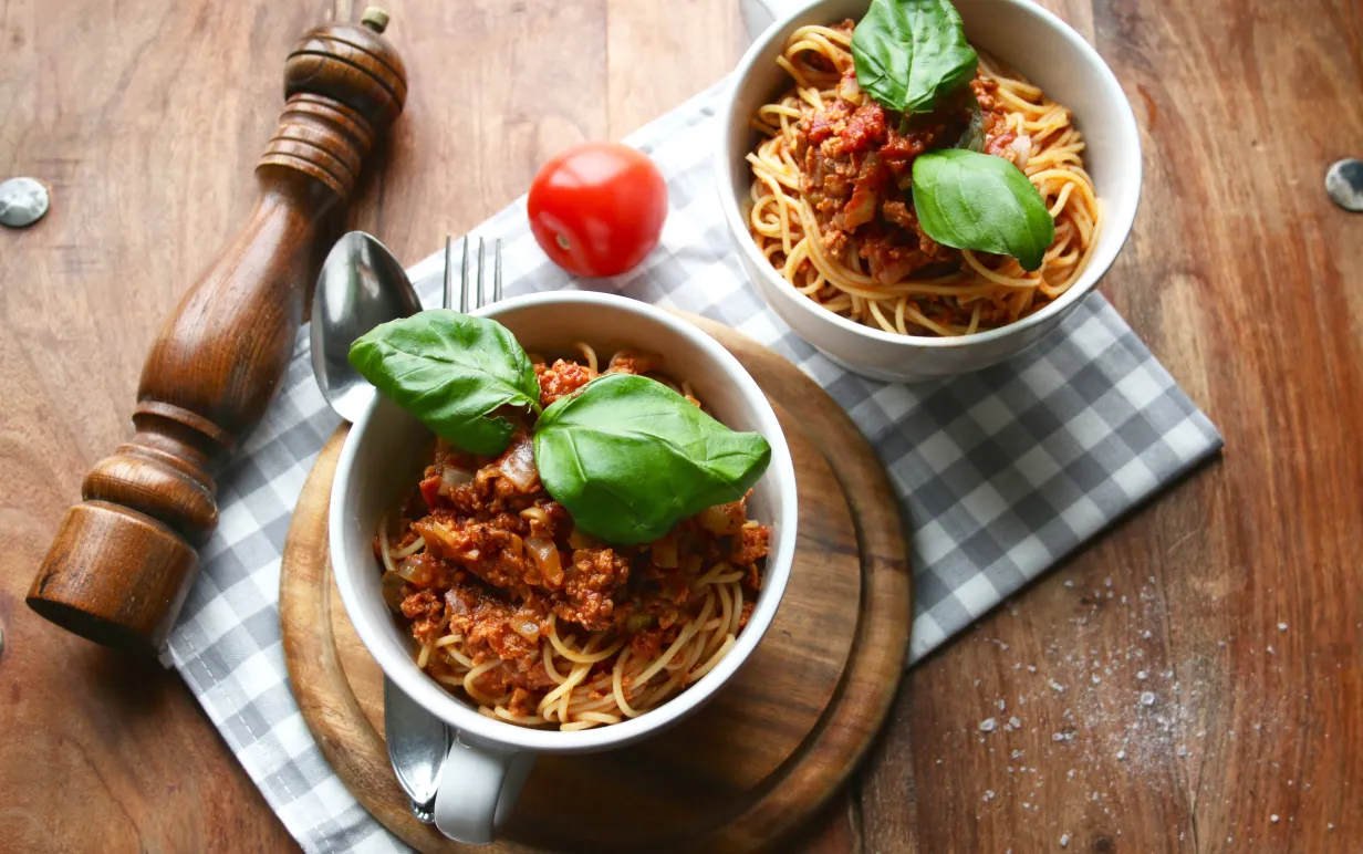 Två skålar med pasta bolognese på ett träbord. Det ligger en pepparkvarn och en tomat brevid skålarna.