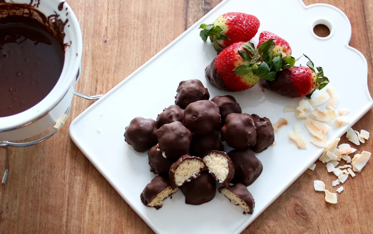 Chokladbitar med kokosfyllning på en vit skärbräda, bredvid ligger några jordgubbar och kokosbitar