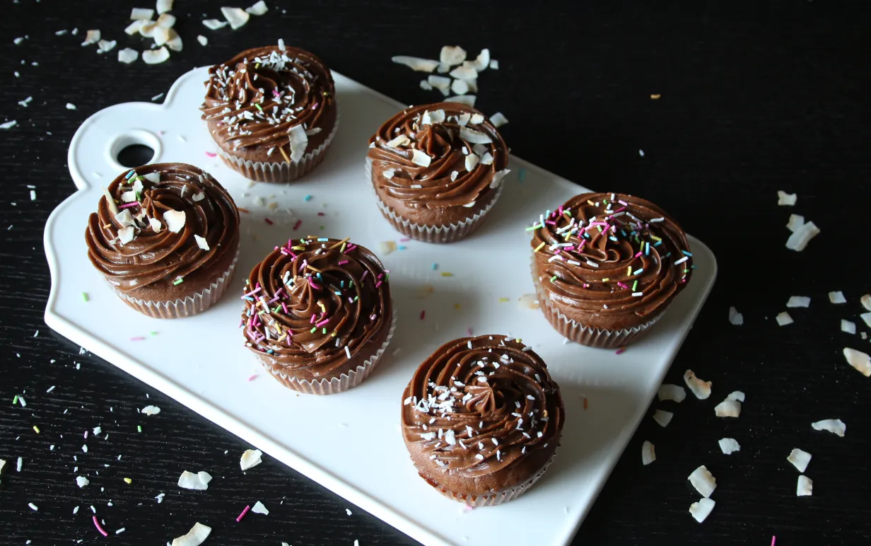 Sex stycken kolamuffins med mockaglasyr, kokos och strössel på en vit skärbräda på ett svart bord.