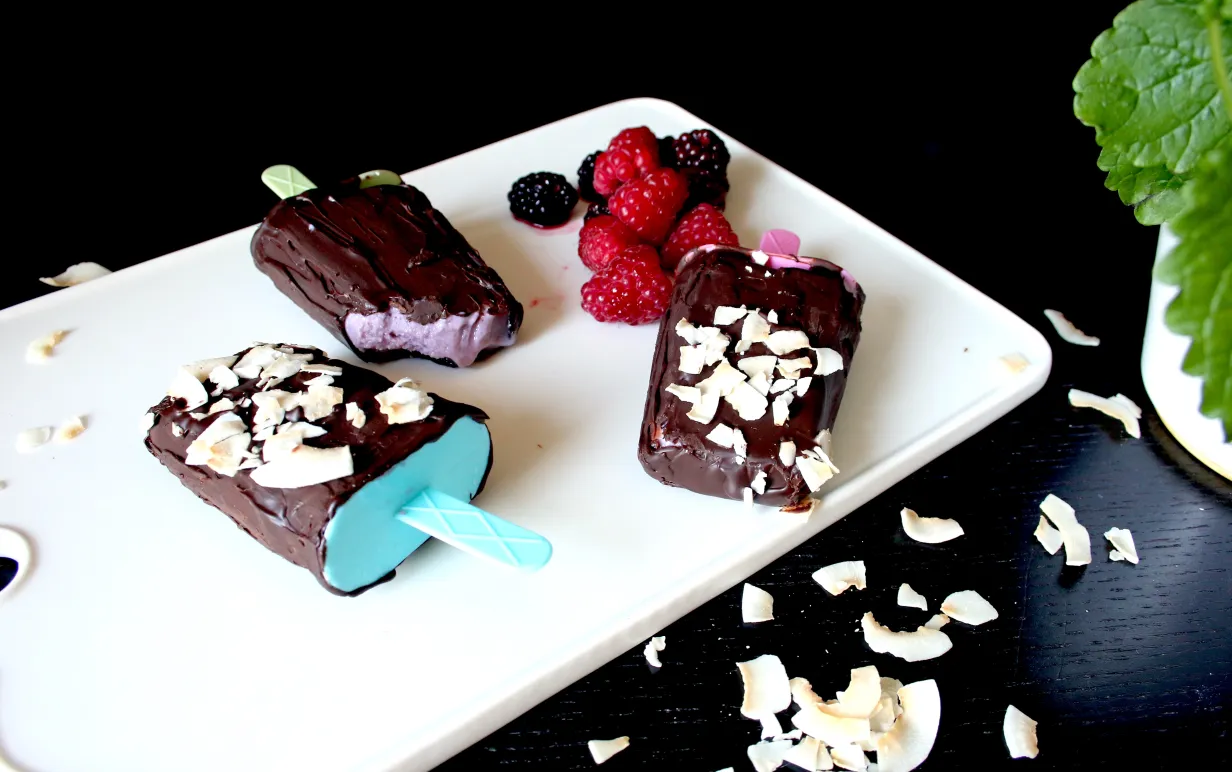 Tre chokladdoppade soygurtglassar på en vit skärbräda, bredvid glassarna ligger några hallon och björnbär