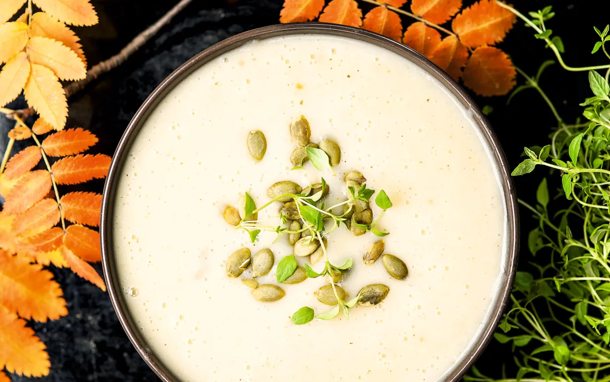 Vit soppa i skål toppad med pumpafrön omgiven av höstlöv