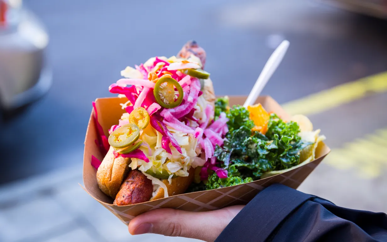 Hand håller upp bröd med vegansk fyllnig och diverse grönsaker. Står på en gata.