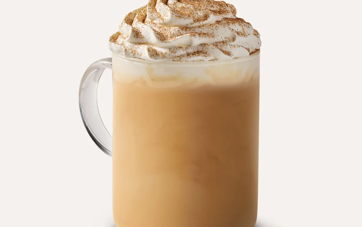 En glaskopp med pumpkin spice latte i och grädde toppad med kanel på toppen mot en vit bakgrund.