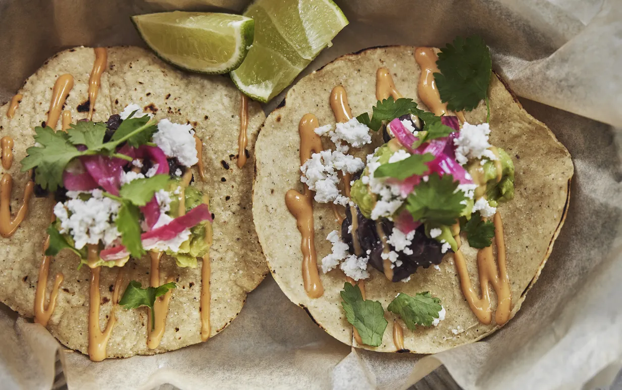 Två, små tacobröd fyllda med bl.a. rödlök, avocado, koriander, vegansk ost och sås, liggandes på bakplåtspapper bredvid limeklyftor.