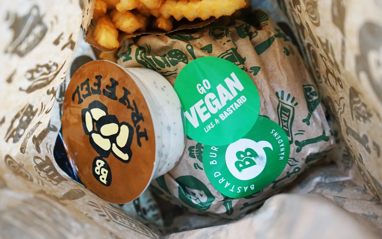 Burgare, pommes och sås från Bastard Burgers i papperspåse. På burgaren finns ett klistermärke med texten "Go vegan like a bastard".