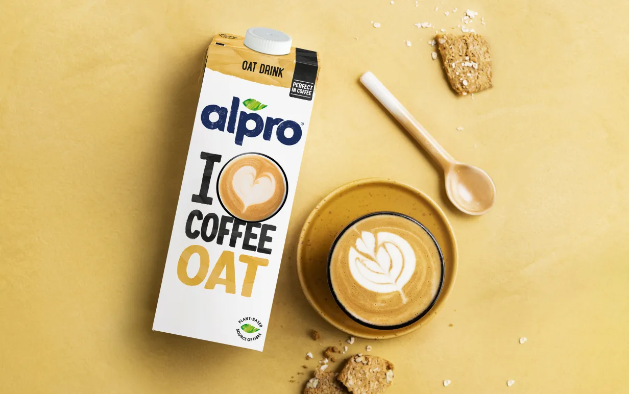 Förpackning med växtbaserad dryck bredvid en kopp latteart på ett gult bord