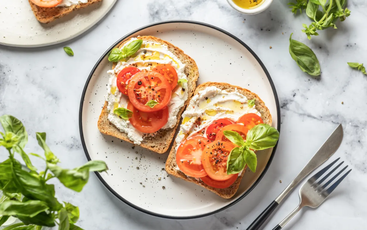 En tallrik med två smörgåsar med vegansk cream cheese och tomater, toppade med basilika
