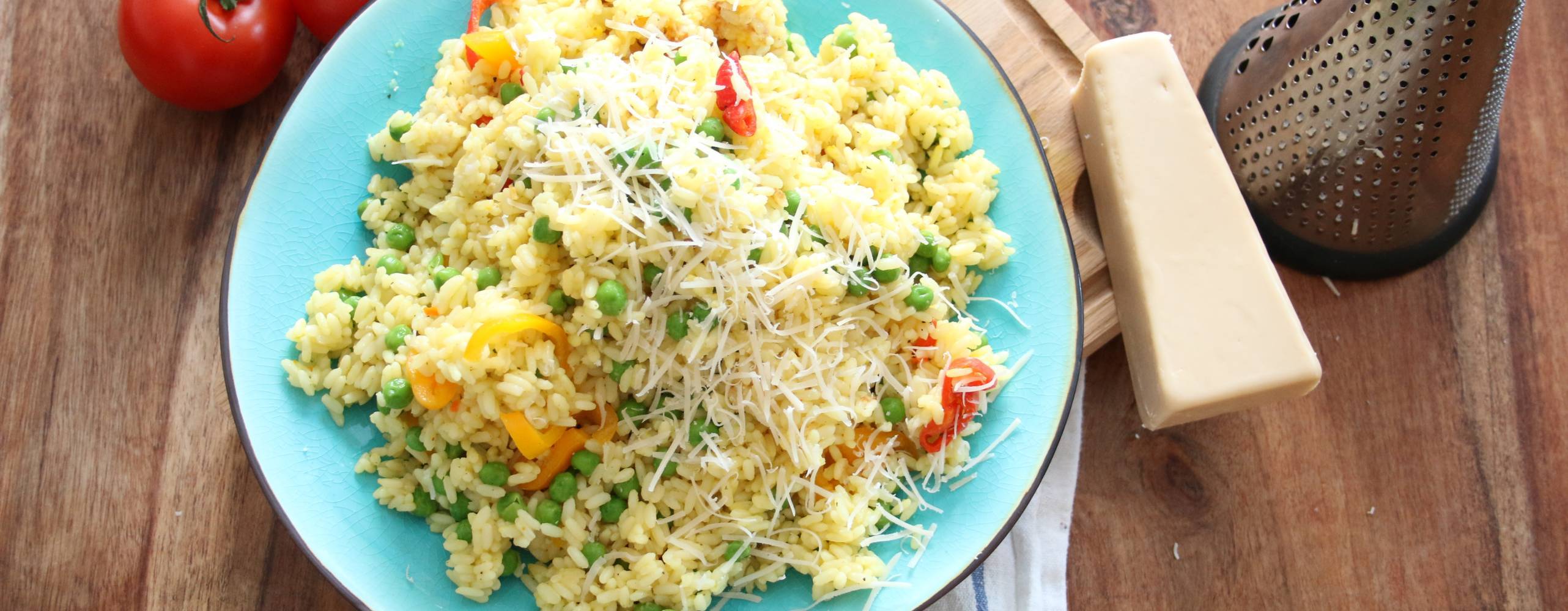 Risotto med gröna ärtor på en turkos tallrik, bredvid tallriken ligger en vegansk parmesanost och några tomater