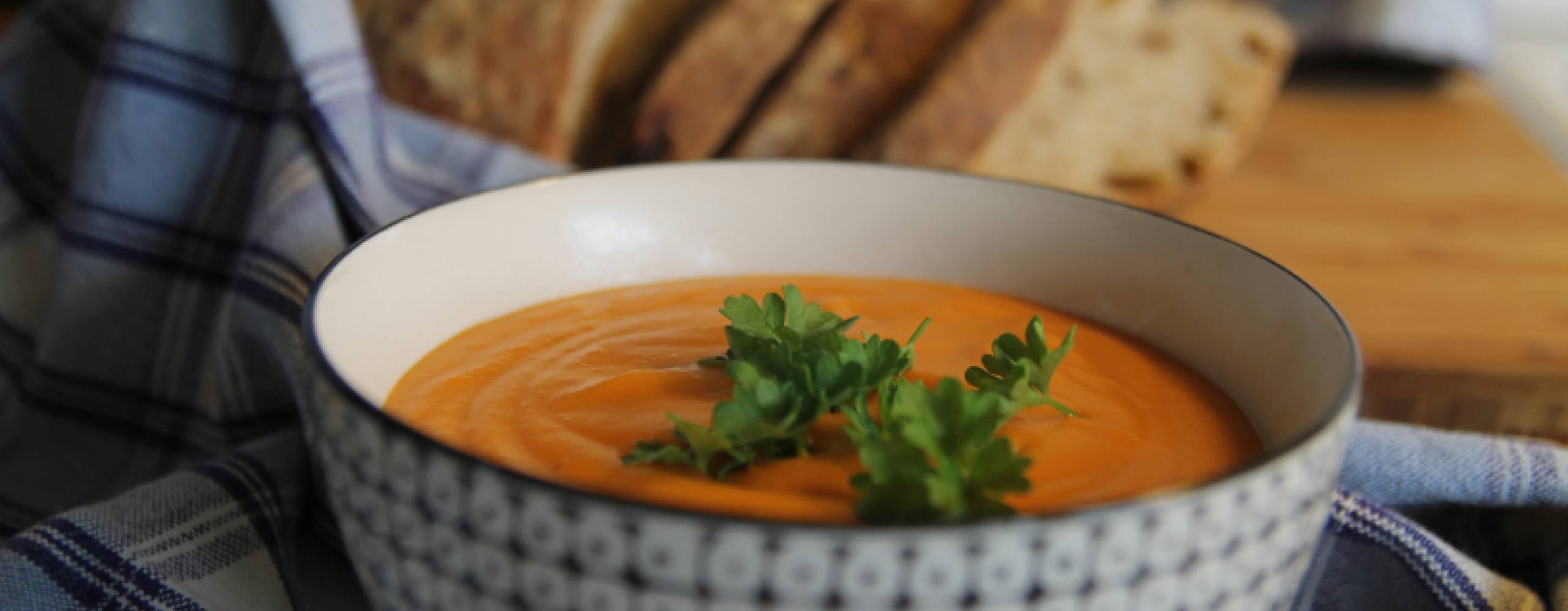 Sötpotatissoppa i en skål på en rutig handduk på ett bord. Det ligger bröd bredvid handduken.