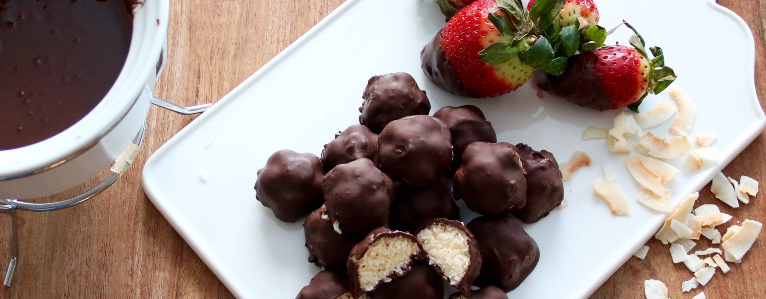 Chokladbitar med kokosfyllning på en vit skärbräda, bredvid ligger några jordgubbar och kokosbitar