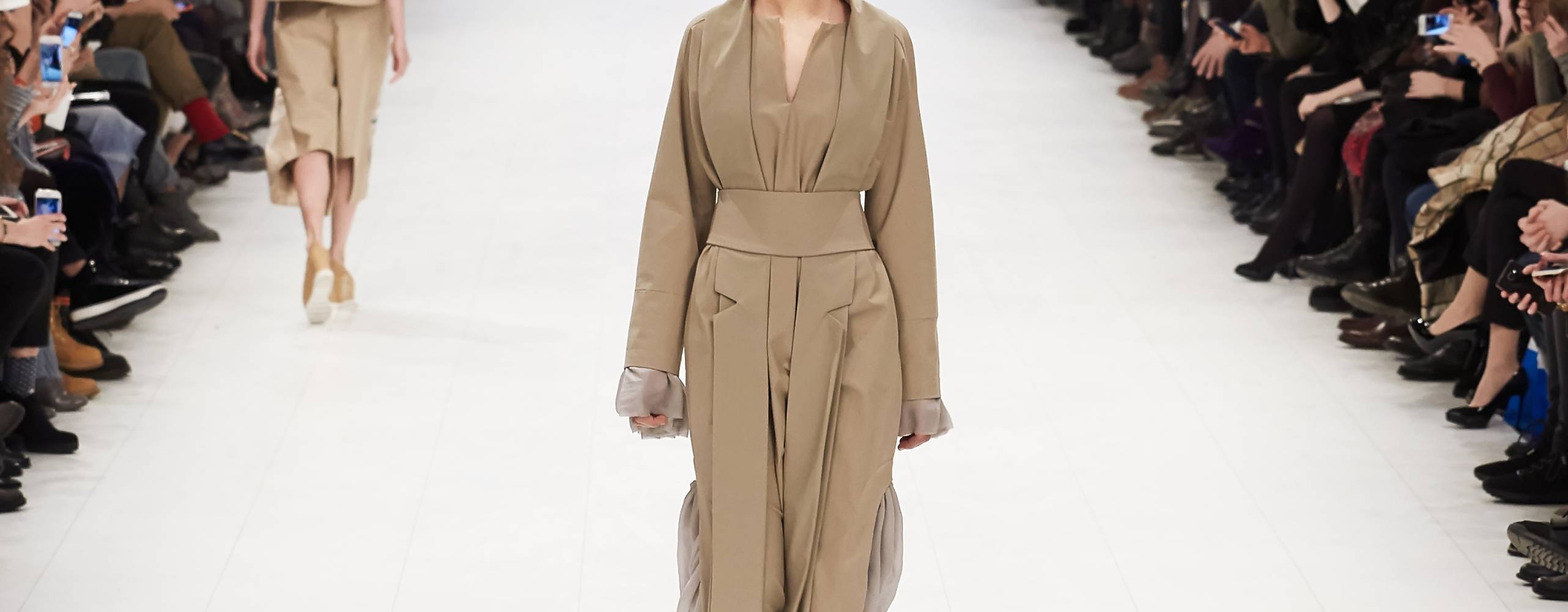 En catwalk med en person i mitten med en trenchcoat och åskådare på varje sida