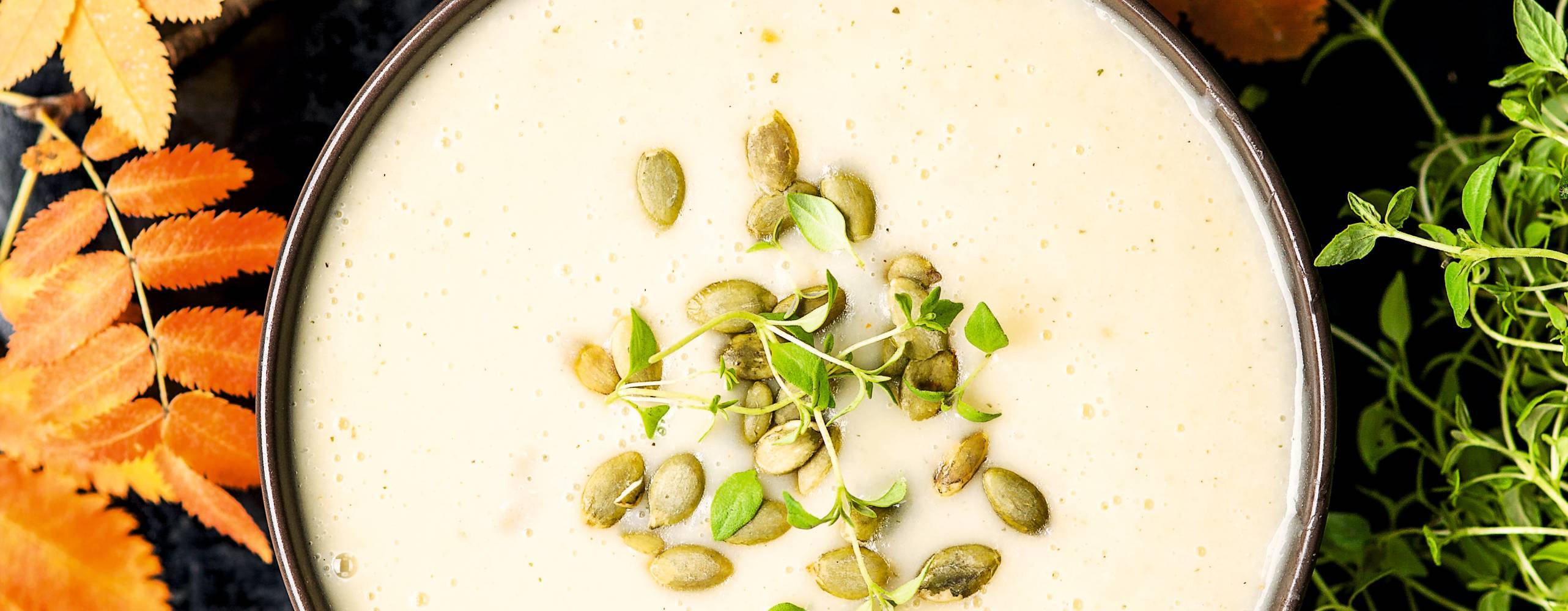 Vit soppa i skål toppad med pumpafrön omgiven av höstlöv