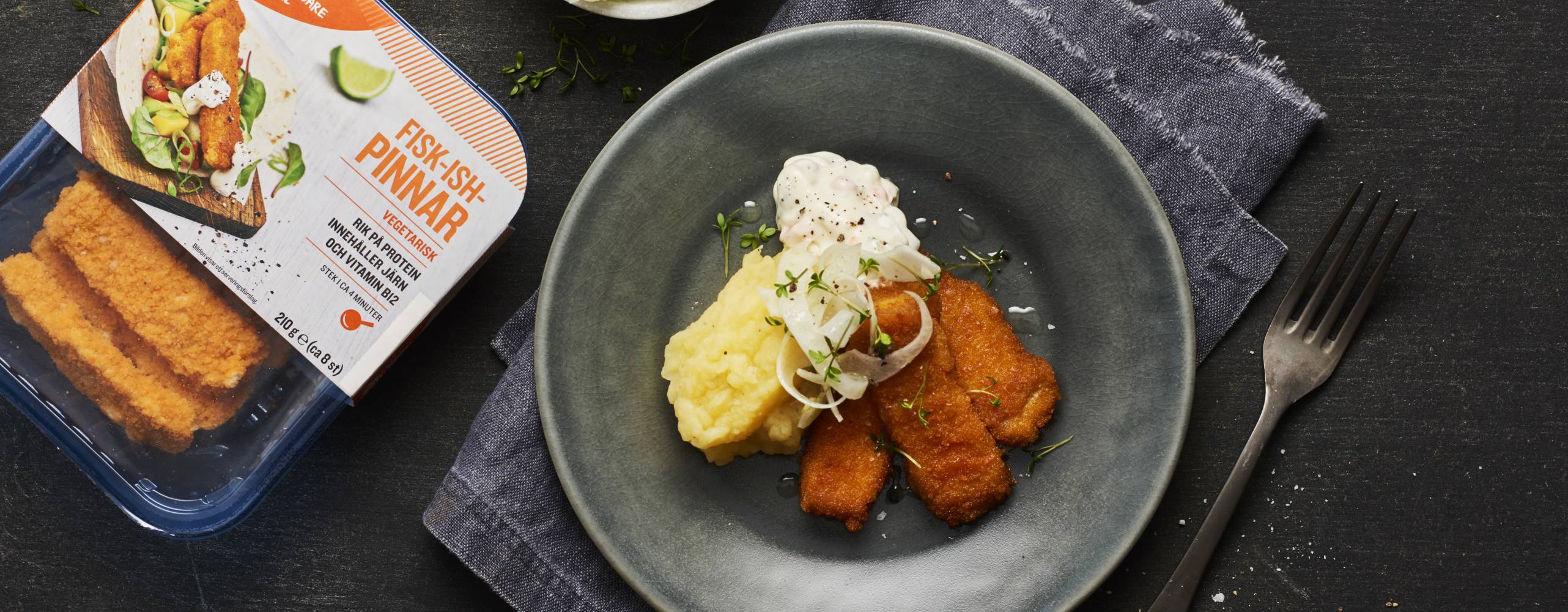 Svart tallrik med veganska fiskpinnar, potatismos och sås bredvid en förpackning veganska fiskpinnar.