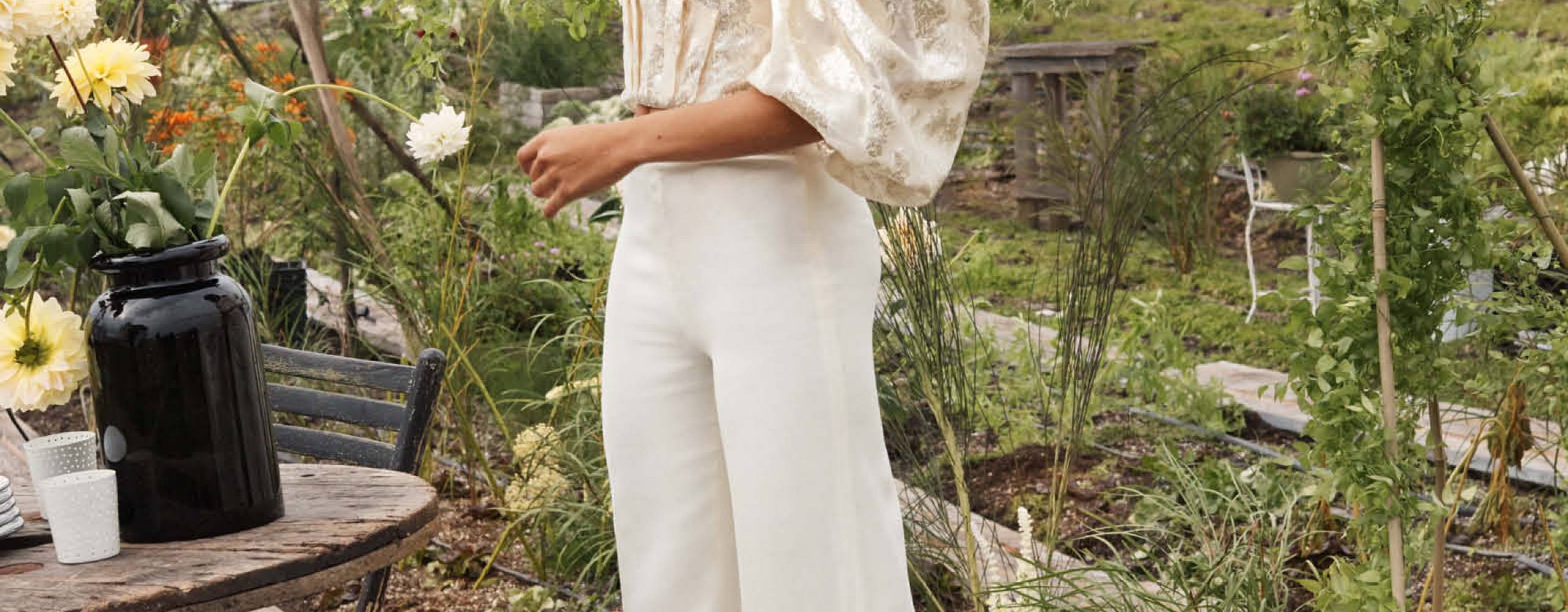 Person med vita byxor och vit topp står i en trädgård.