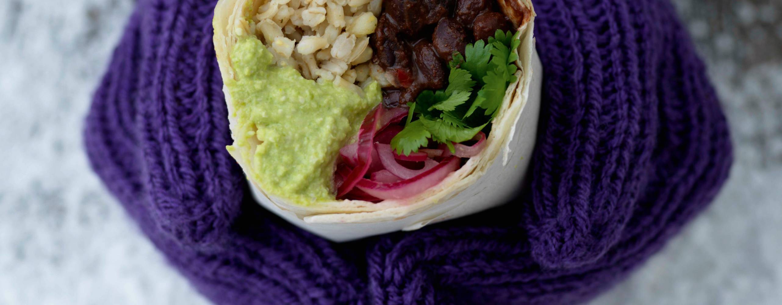 händer med lila vantar håller i en burrrito med krämiga svarta bönor, guacamole och ris