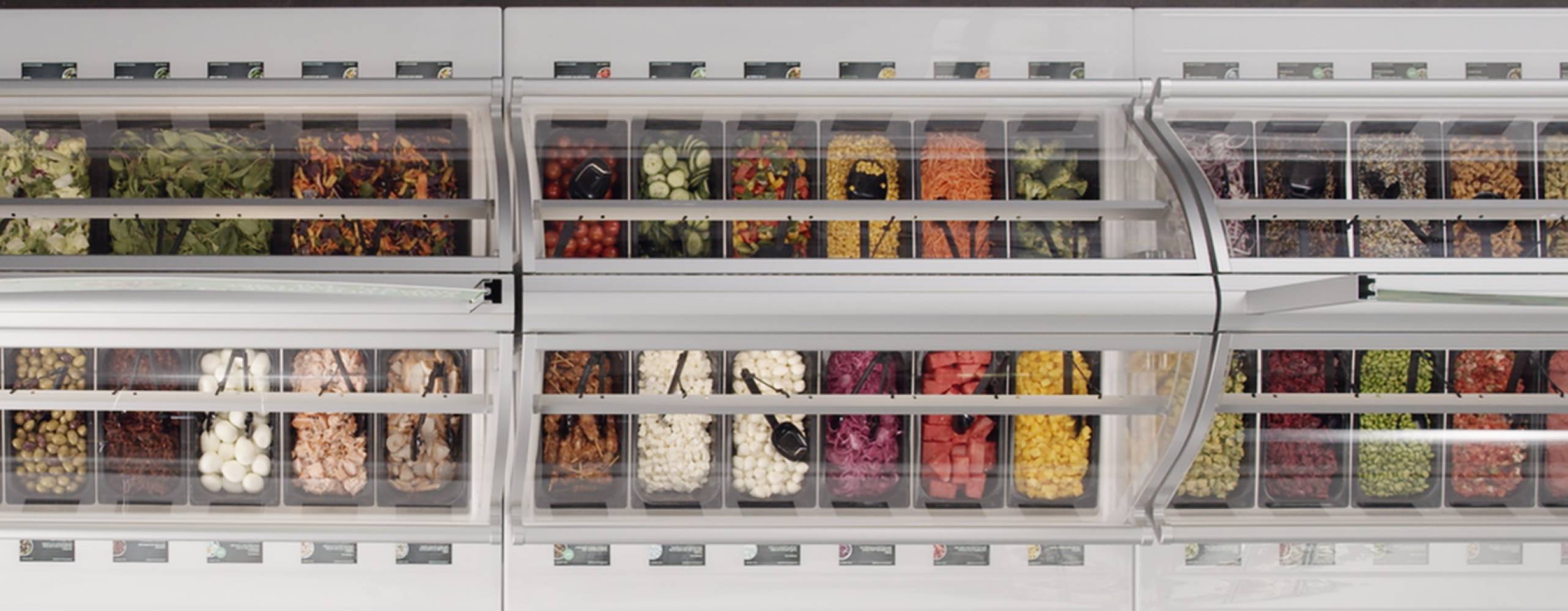 Salladsbar med många olika, färgglada alternativ, fotad ovanifrån mot mörkgrått golv.