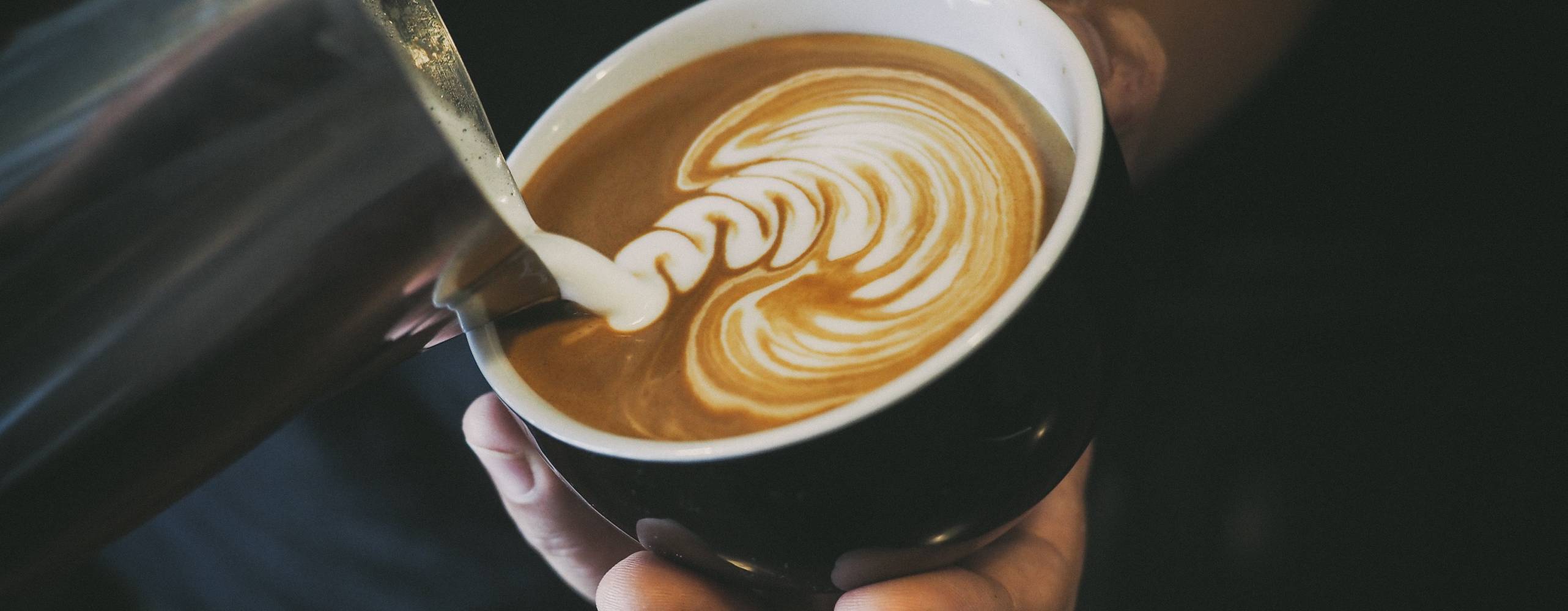 "Mjölk" hälls i kopp med kaffe. Koppen hålls i en hand i dovt belyst miljö.