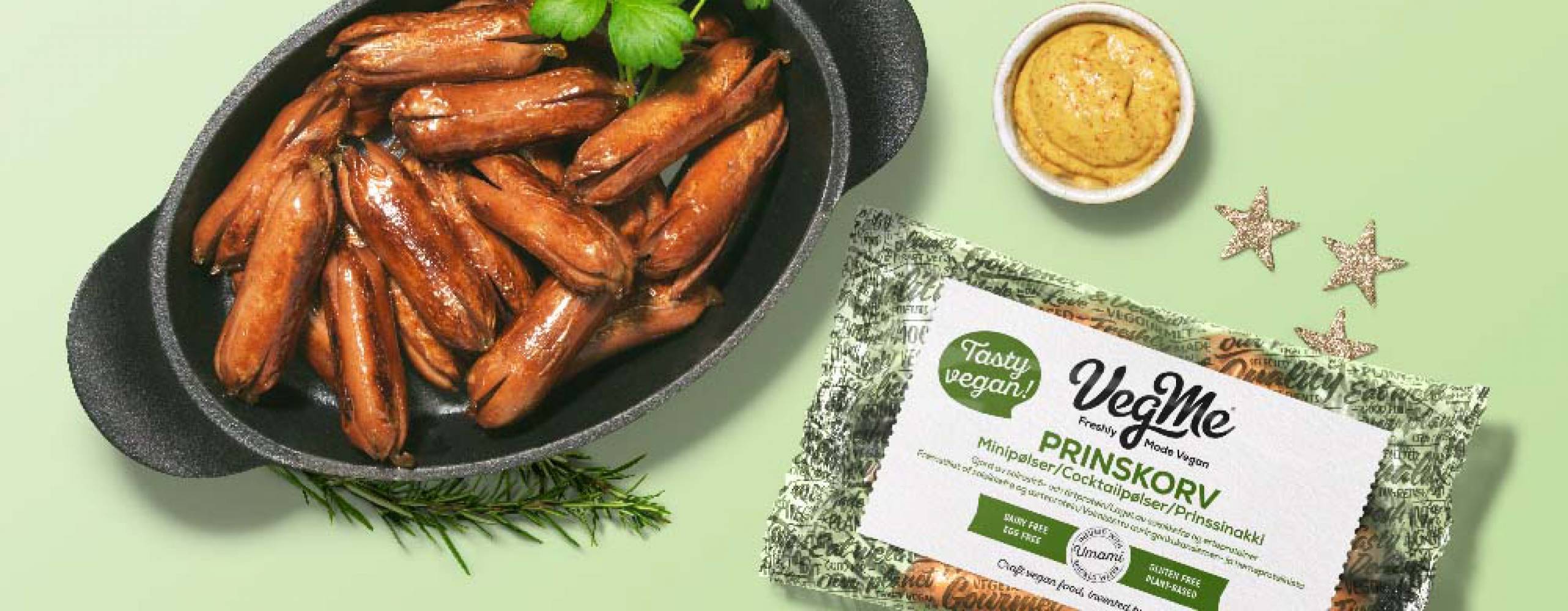 Prinskorv från VegMe i gjutjärnsgryta mot grönbakgrund, med förpackning och en liten skål senap bredvid.