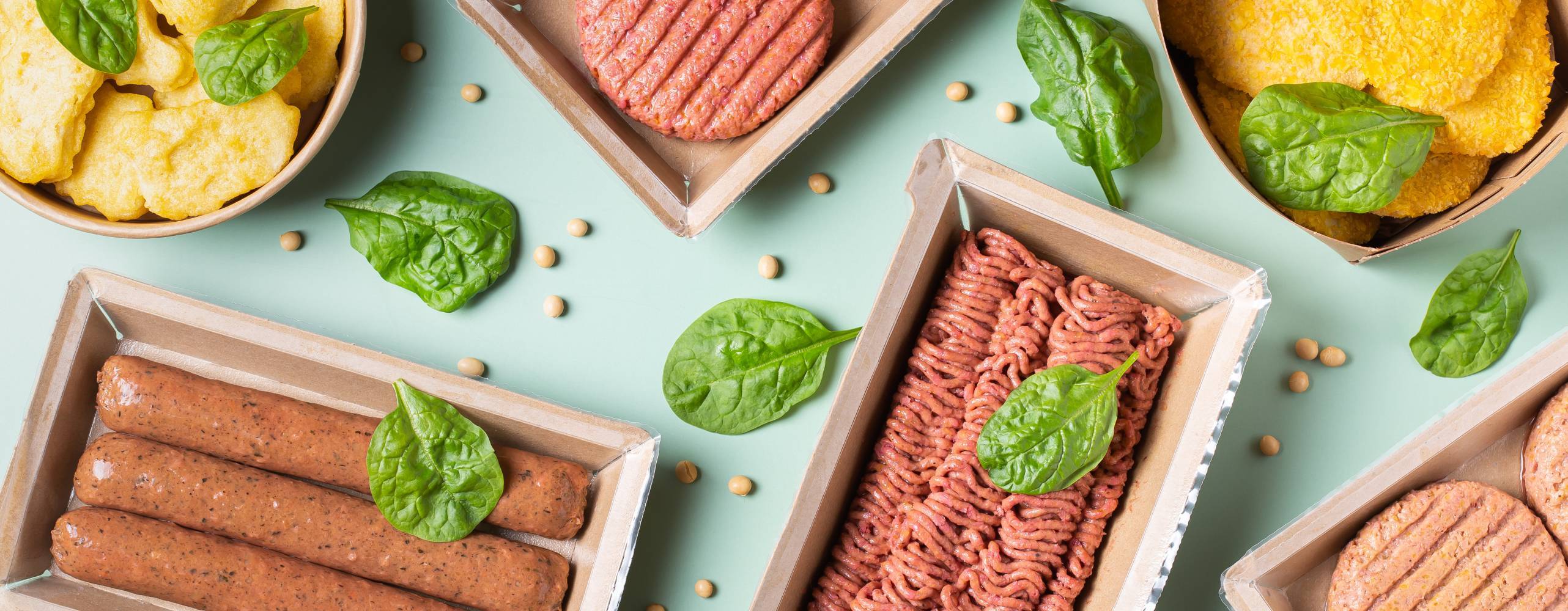 Flera olika växtbaserade köttsubstitut upplagda på ett mintgrönt bord med basilikablad utspridda.
