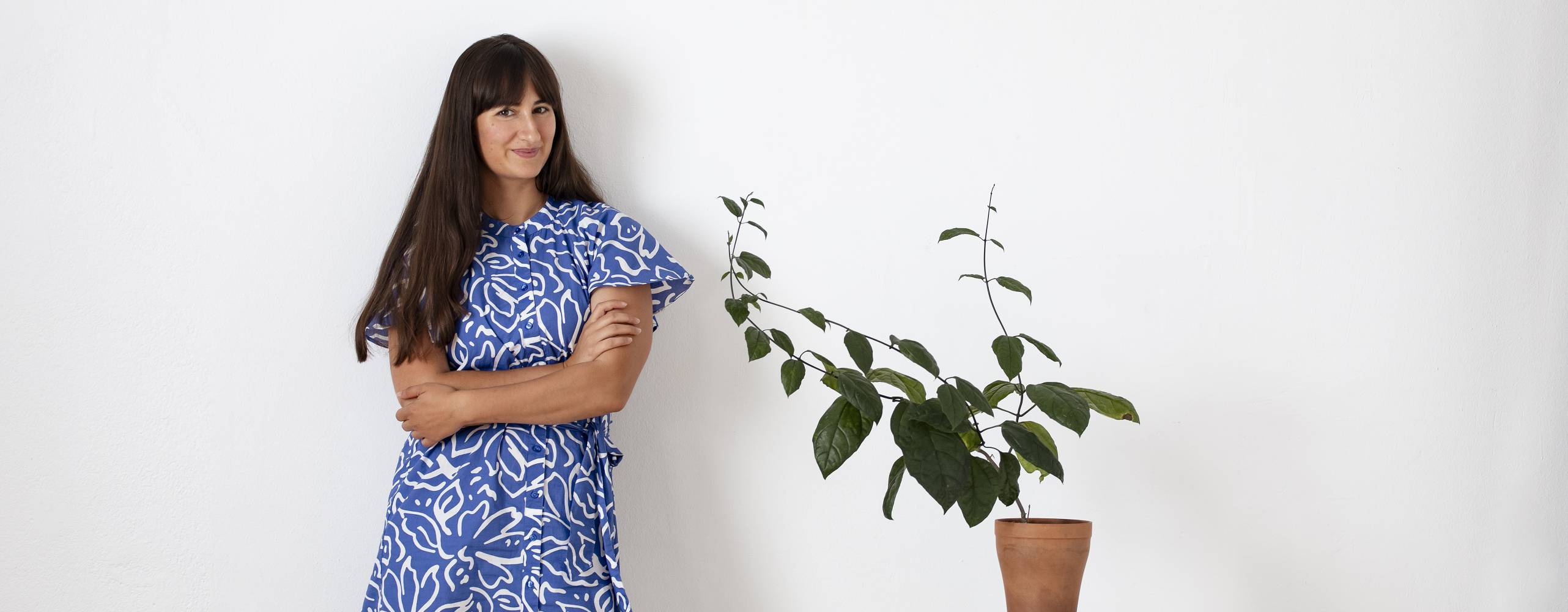 Mörkhårig kvinna i blå klänning bredvid en grön krukväxt mot en vit vägg.