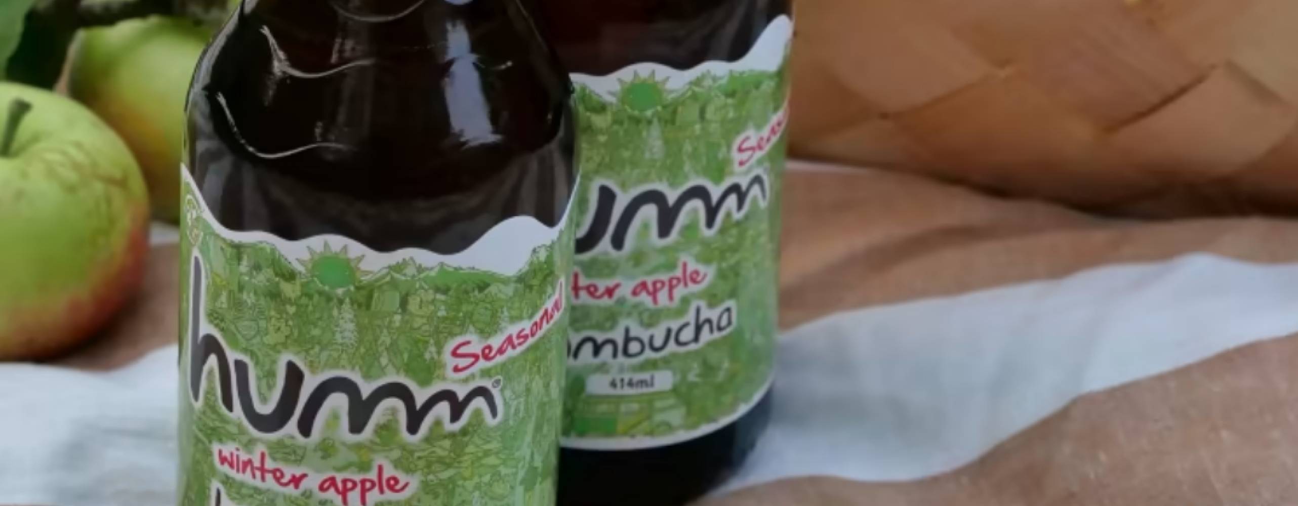 Två glasflaskor med kombutcha på en filt med en korg och äpplen i gakgrunden.