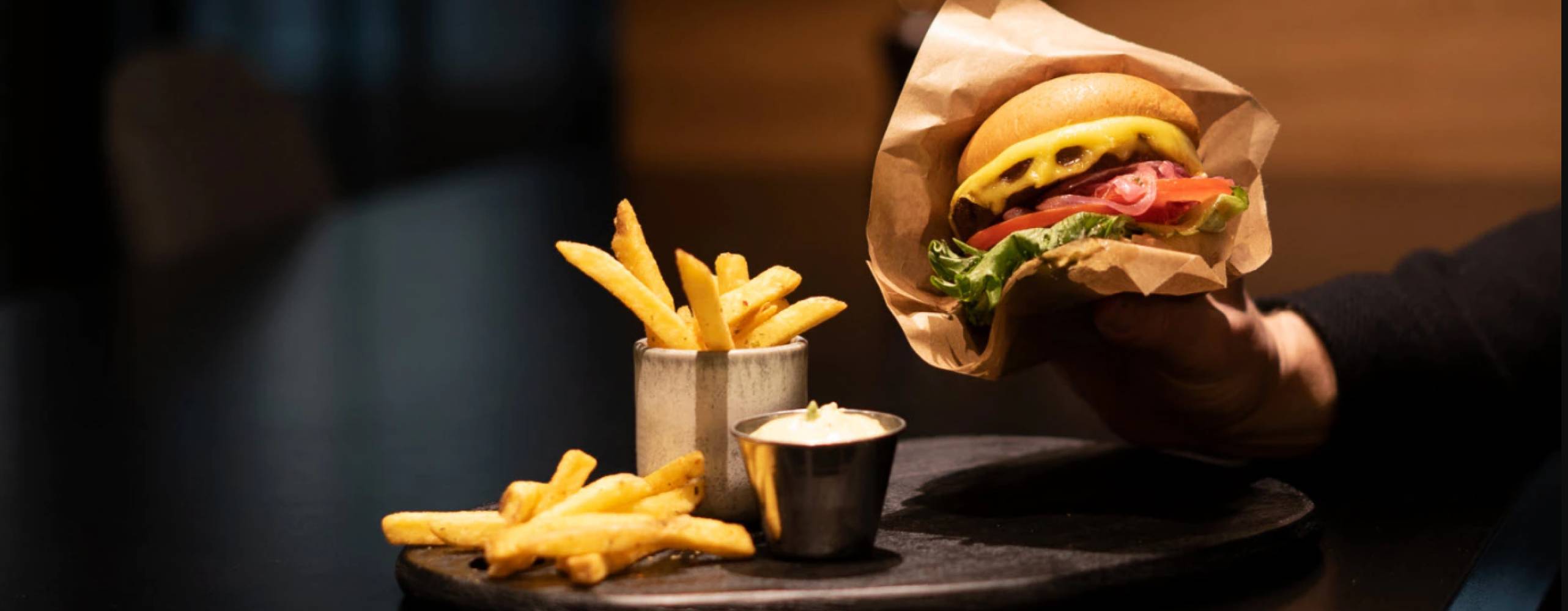 en hand som håller i en vegoburgare med pommes och dip på ett bord i en mörk restaurangmiljö