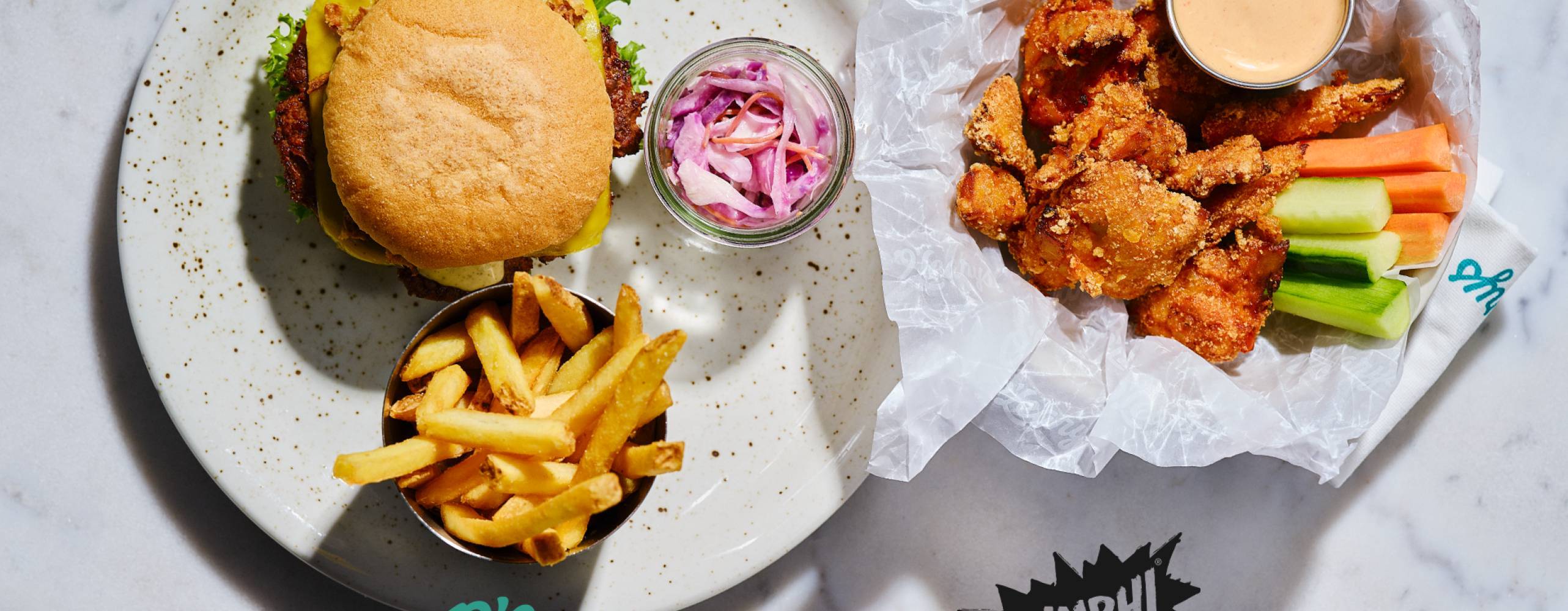 En tallrik med hamburgare, pommes och coleslaw och en med veganska chicken wings, grönsaker och dip. Bild tagen uppifrån.