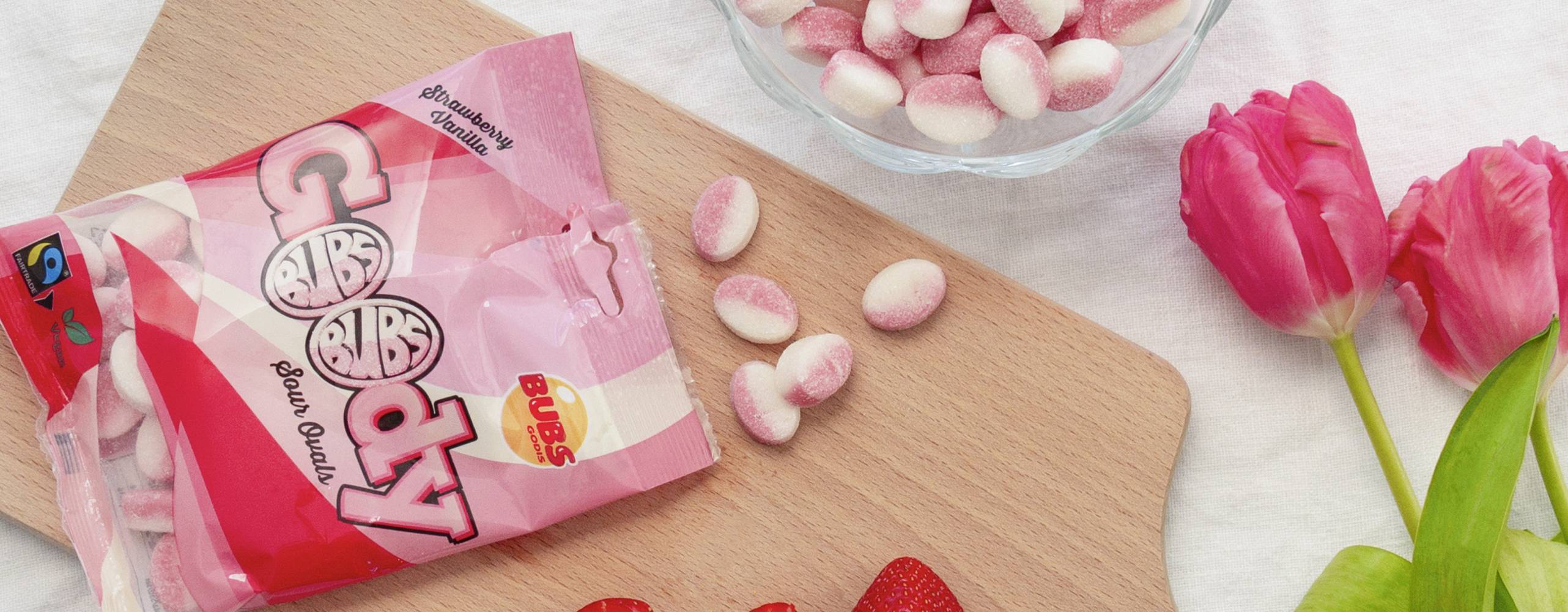 En påse godis öppnat och utspridd på en skärbräda i trä med jordgubbar och röda tulpaner runt