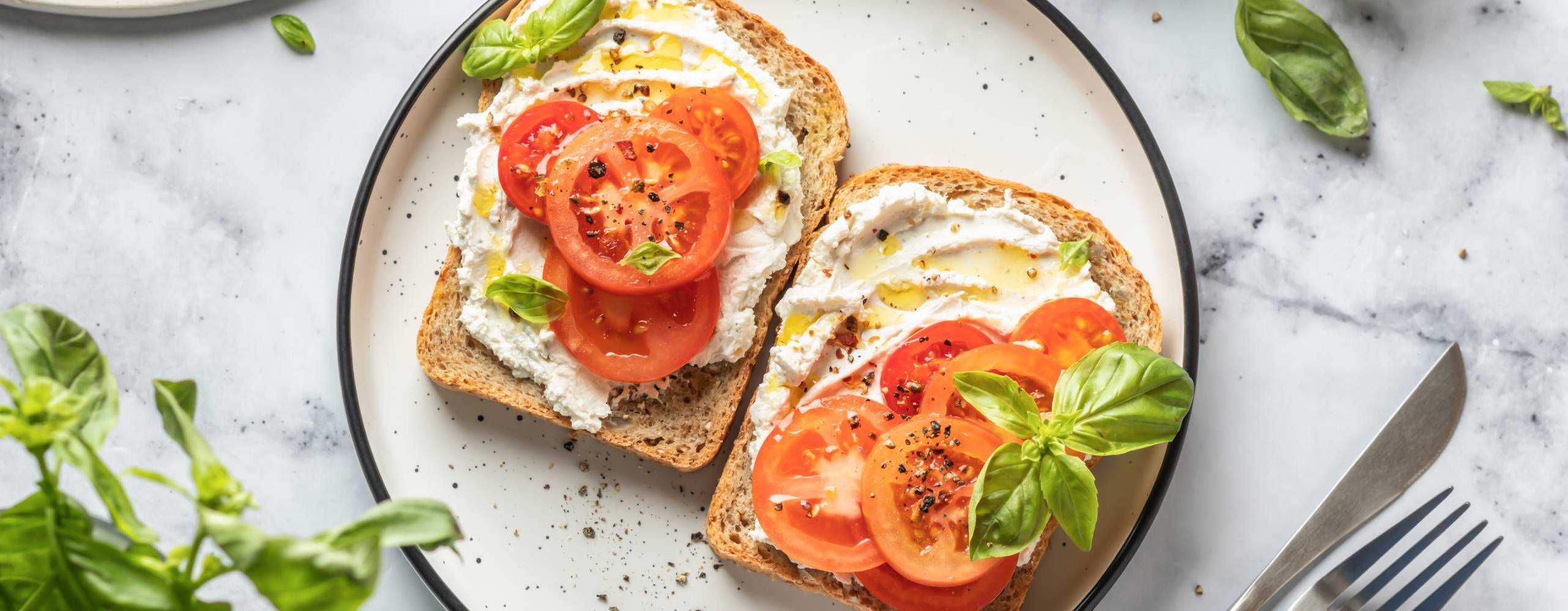 En tallrik med två smörgåsar med vegansk cream cheese och tomater, toppade med basilika