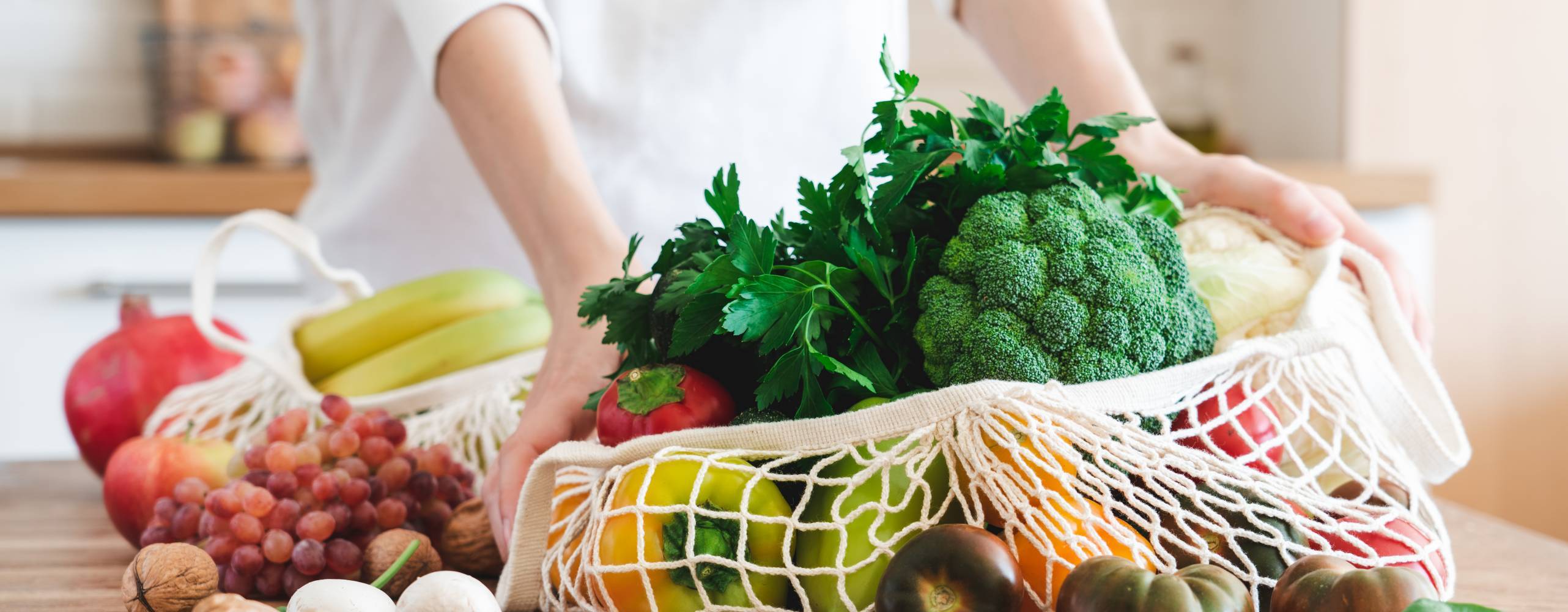 Fruits and vegetables in cotton reusable net mesh bag on wooden table in modern bright kitchen. Organic vegan food set. Sustainable lifestyle concept